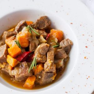 Beef & Butternut Squash Stew in a white bowl with the edge sprinkle with a red spice