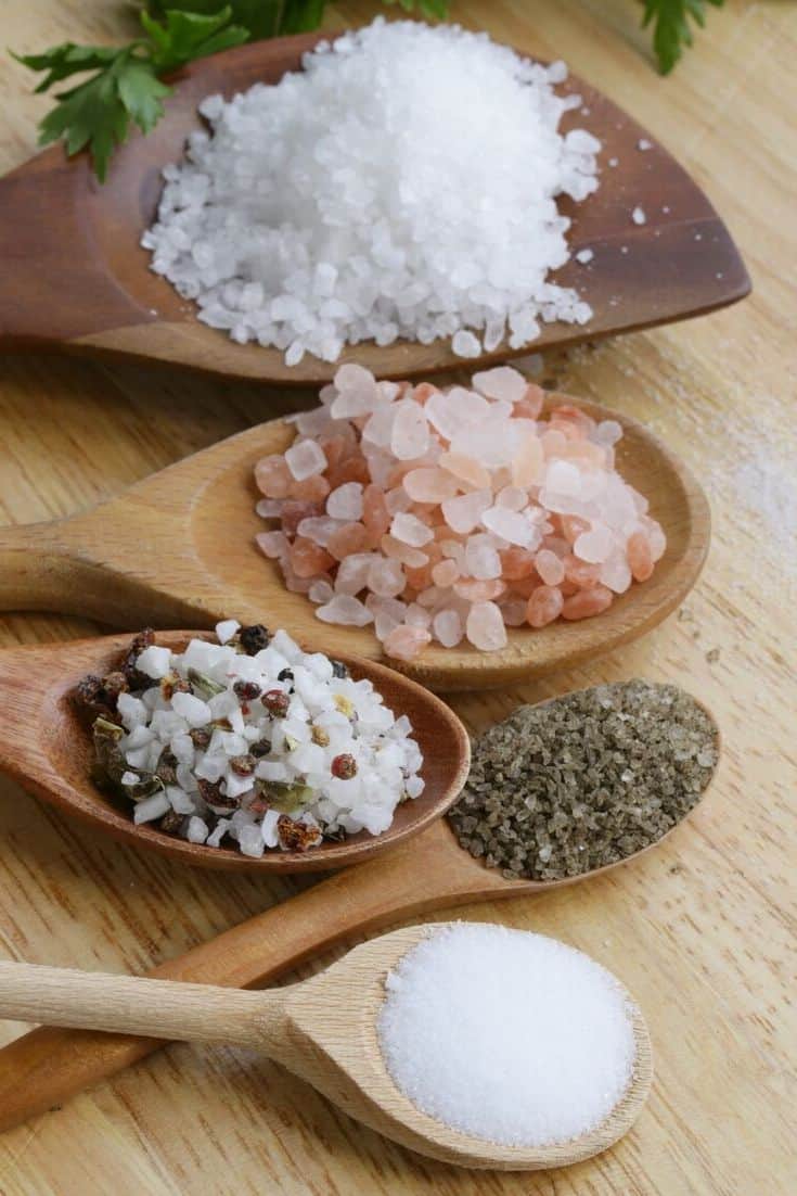 Different types of salt in various sized wooden spoons on a wood surface