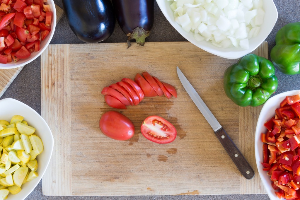 Prepping fresh vegetables for cooking in the kitchen with diced zucchini, tomato, white onion , green capsicum or bell pepper and eggplant arranged around a wooden chopping board with a knife